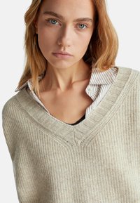 Beige pletený svetr s designem do V, se strukturovaným žebrovaným vzorem a pod ním vrstvenou košilí s proužky.