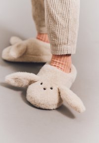 Tan plüsskaninulpantoflar med långa öron, svarta prickar som ögon och rundad form. Bärs tillsammans med ljus korallrandiga strumpor och ribbade beige byxor.