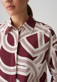 Camicia di cotone con un motivo geometrico bordeaux e crema, dotata di colletto a punta e chiusura con bottoni. Texture morbida con linee distintive.