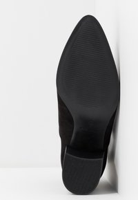 Svart spetsig sko med en sål av gummi med räfflor och låg klack, placerad upprätt mot en vit hörnyta.