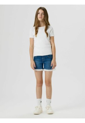 Mladá dívka stojí, má na sobě bílou košili s krátkým rukávem, džínové šortky s krajkovou výšivkou, bílé ponožky a bílé tenisky, neutrální pozadí.