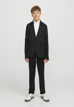 Αγόρι στέκεται, φορώντας μαύρο κοστούμι με λευκό πουκάμισο και λευκά αθλητικά, μπροστά σε απλό φωτεινό φόντο.