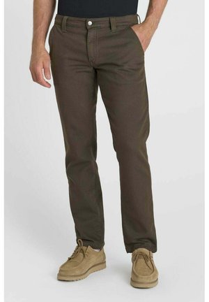 Uomo che indossa pantaloni marrone scuro e scarpe stringate marrone chiaro, in piedi con una mano in tasca su sfondo semplice.