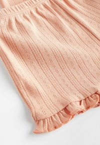 Țesătura de tricot piersică cu un model texturat ondulat, având un brâu strâns și un detaliu cu volan la partea de jos.