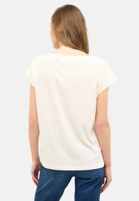 Cremefarbenes T-Shirt mit kurzen Ärmeln, lockerer Passform, runder Ausschnitt und weichem Stoff. Trägt es mit blauen Jeans, von hinten betrachtet.