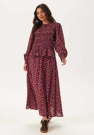 Kvinde iført en langærmet burgunderrød kjole med hvide polkaprikker og brune sneakers, stående mod en ensartet lys baggrund.