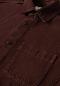 Camicia di cotone marrone con colletto button-down, bottoni neri e taschino sul petto con dettagli di cucitura visibili. Texture morbida.
