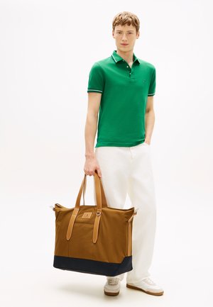 Beige en marineblauwe tas met leren handvatten, met een rechthoekige vorm en een logo-label, gedragen door een persoon in casual kleding.