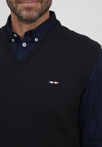 Granatowa dzianinowa kamizelka z dekoltem w serek, z haftowanym poziomym paskiem w kolorach czerwonym, białym i niebieskim po lewej stronie klatki piersiowej.