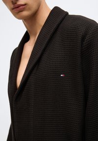 Melns austs halāts ar šalli apkakli, teksturētu audumu un mazu izšūtu logotipu sarkanā, baltā un zilā krāsā.