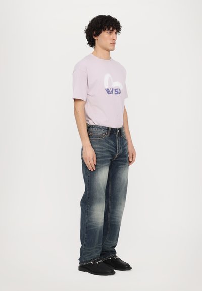 T-shirt violet clair avec un logo style graffiti, associé à un jean en denim bleu foncé avec un design effacé et des chaussures noires.