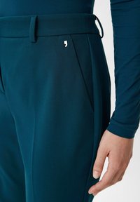 Ciemnoniebieskie spodnie z teksturowanej tkaniny, z kieszenią boczną i małym srebrnym logo. Pas jest gładki i dopasowany.