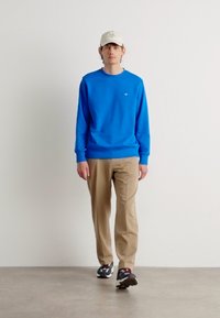 Blauwe sweatshirt, beige broek en zwart-witte sneakers. Model draagt een crèmekleurige pet. Eenvoudige achtergrond, staande pose.