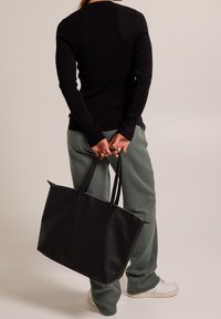 Person som bär en svart långärmad tröja och grå byxor, håller en stor svart axelväska bakom ryggen, står på vita sneakers.