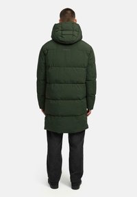 Cappotto imbottito verde scuro con cappuccio, caratterizzato da pannelli trapuntati orizzontali e una parte posteriore più lunga. Indossato sopra pantaloni scuri dalla texture e scarpe nere.