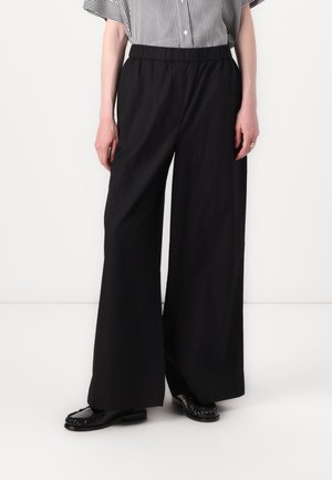 Personne portant un pantalon noir large et ample, des ballerines noires, et une chemise rayée noire et blanche, debout devant un fond blanc uni.