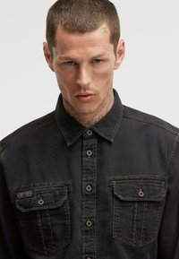 Schwarzes Denim-Hemd mit Knopfleiste, zwei aufgesetzten Taschen vorne, sichtbaren Nähten und Kragen. Der Stoff hat eine strukturierte Optik.