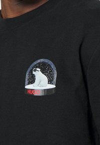 Sudadera negra con un pequeño gráfico de un oso polar dentro de una bola de nieve. La bola tiene una cúpula azul y copos de nieve blancos, con "HUGO" en rojo debajo.