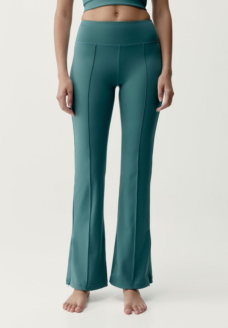 Vrouw in tealkleurige, hooggetailleerde, uitlopende legging met naaddetails, staat op blote voeten voor een effen witte achtergrond.