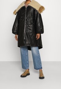 Cappotto oversize in ecopelle nera con collo in pelliccia beige, dotato di zipper frontale, vita con cordino e tasche ampie, abbinato a jeans blu.