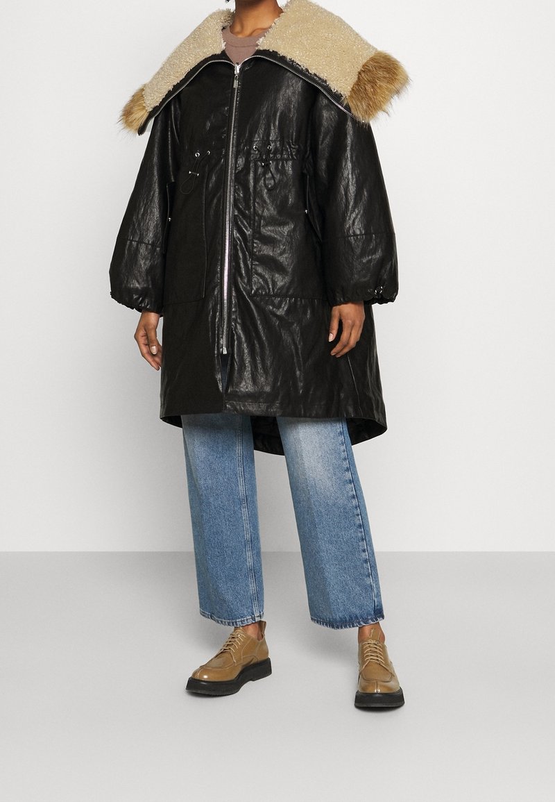 Cappotto oversize in ecopelle nera con collo in pelliccia beige, dotato di zipper frontale, vita con cordino e tasche ampie, abbinato a jeans blu.