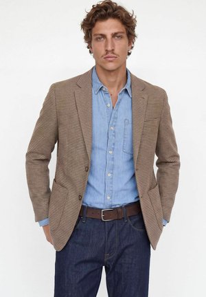 Un mannequin porte un blazer marron à motifs sur une chemise bleue, avec un jean foncé et une ceinture marron, devant un fond blanc uni.