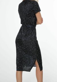 Svart klänning med en figurnära midja, korta ärmar och en slits på sidan. Har ett mönster av vita prickar på ett texturerat tyg.