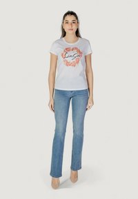 Weißes Baumwoll-T-Shirt mit einem floralem Design in Rot und Orange sowie schwarzem Text in der Mitte, kombiniert mit hellblauen Jeans und beigen hohen Schuhen.