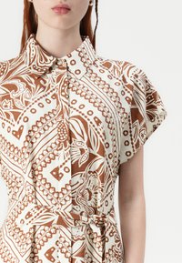 Abito camicia a maniche corte con motivo marrone e bianco, colletto e dettaglio legato in vita. Presenta disegni floreali e geometrici.