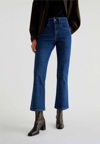 Blue high-waisted flared jeans made of denim, featuring a five-pocket design and subtle stitching details. Worn with brown block-heeled boots.