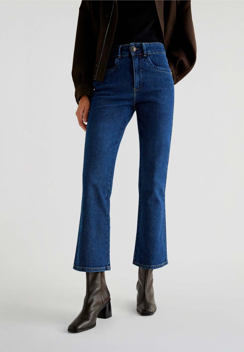 Blue high-waisted flared jeans made of denim, featuring a five-pocket design and subtle stitching details. Worn with brown block-heeled boots.