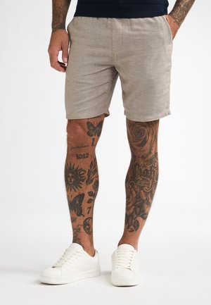 Light beige linen shorts with an elastic waistband and side pockets, paired with white sneakers. Visible tattoo details on legs.