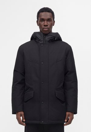 Jeune homme portant un manteau d'hiver noir à capuche avec boutons sur le devant et poches à rabat, debout devant un fond clair uni.