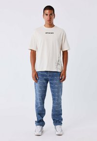 Camiseta de manga corta color crema con el logo negro "PXP" y un gráfico multicolor en el lateral, combinada con unos jeans azules que presentan un patrón de texto repetido.