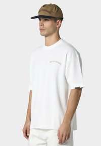 Hvid kortærmet T-shirt med løs pasform, der har en subtil broderet tekst "PAS DE MONACO" i brun. Sammen med en brun kasket med en sort skygge.
