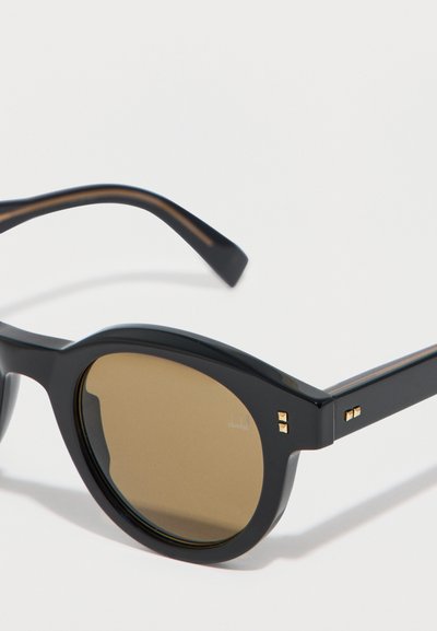 Lunettes de soleil rondes noires avec des verres teintés marron et de petits accents carrés dorés aux coins de la monture et sur les branches.