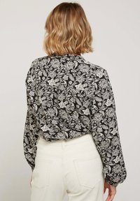 Blouse noire avec motif floral blanc, manches longues et col à boutons. Portée avec un pantalon taille haute de couleur claire.
