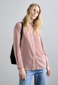 Camicia a righe rosse e bianche con colletto, maniche lunghe e taglio morbido, abbinata a jeans di denim azzurri e una borsa nera.