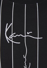 Schwarzer Stoff mit vertikalen weißen Streifen und einem weißen, gestickten abstrakten Liniendesign, das einer Signatur oder stilisiertem Text ähnelt.