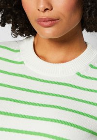 Weißer Pullover mit grünen horizontalen Streifen. Gerippter Kragen und Bündchen. Weiche Textur, Nahaufnahme mit erkennbaren Hauttönen im Hintergrund.