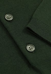 Sötétzöld kötött pulóver, gallérral, bordás mandzsettákkal és két fémgombbal, amelyek kör alakú perforációs mintával rendelkeznek.
