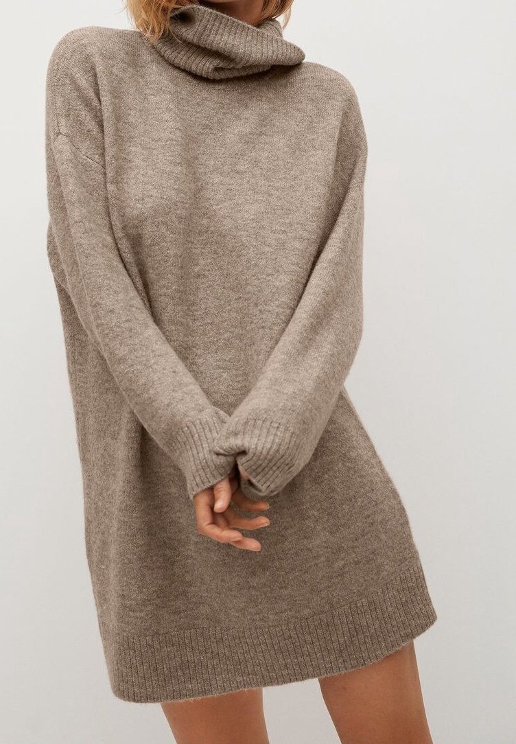 Femme portant une robe-pull à col roulé longue et ample de couleur taupe avec des poignets et un ourlet côtelés, les mains jointes devant.