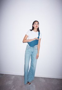 Borsa a tracolla in denim indossata sul petto, abbinata a jeans larghi di colore blu chiaro e una t-shirt attillata bianca su uno sfondo neutro.