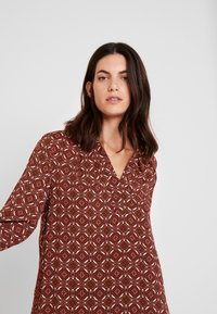 Blusa com padrão vermelho, apresentando um decote em V e mangas longas, com designs geométricos em creme e tons de laranja sobre um tecido texturizado.
