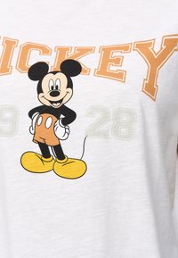 Valkoinen lyhythihainen T-paita, jossa on kuvasto sarjakuvahahmosta, jolla on mustat korvat, oranssit shortsit ja keltaiset kengät. Teksti kuuluu "MICKEY 1928."