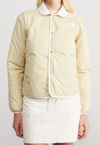 Vaaleankeltainen vuorillinen takki, jossa valkoinen kaulus, painonapit ja ompelu koristeet. Päällä valkoinen denimhame.