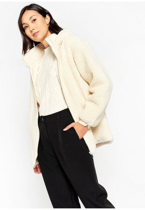 Cremefarbene Kunstfelljacke mit hohem Kragen, Reißverschluss und lockerer Passform, kombiniert mit einem strukturierten Strickpullover und schwarzen Hose.
