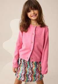 Rosa Wollstrickjacke mit goldenen Knöpfen und Rundhalsausschnitt. Kombiniert mit einem mehrfarbigen Paillettenrock mit wellenförmigen Mustern.