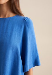 Top in lino blu con collo rotondo, maniche corte e pieghe delicate sulle spalle, caratterizzato da una superficie liscia e texturizzata.