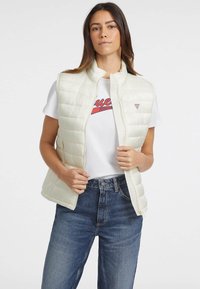 Weißes Daunenwesten mit Reißverschluss, hochgeschlossen und mit Seitentaschen, getragen über einem weißen T-Shirt und blauen Jeans.